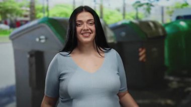 Mavi gömlekli kıvrımlı genç bir kadın telefon jesti yapıyor ve arka planda geri dönüşüm kutuları olan bir şehir caddesinde dışarıyı işaret ediyor..