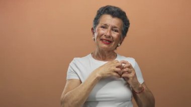 Gri saçlı yaşlı bir kadın, elleri turuncu arka planda, beyaz bir gömlek giymiş, içten gülümseyen, sevgi ve mutluluğu ifade eden bir kalp hareketi yapıyor..