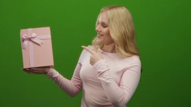 Pembe tişörtlü bir kadın canlı yeşil bir arka planda pembe bir hediye kutusuna bakıp gülümsüyor.