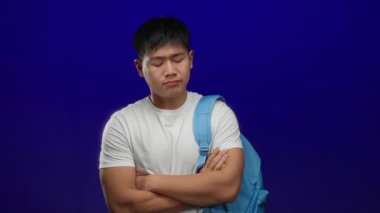 Kolları çapraz mavi sırt çantası takan genç bir adam mavi arka planda izole edilmiş, öğrenci duygularını aktarıyor..