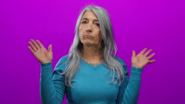 Mavi kazaklı, gri saçlı yaşlı bir kadın, izole edilmiş, pembe bir arka plana karşı poz veriyor. Sorgu jestinde ellerini kaldırarak ifade ediyor..
