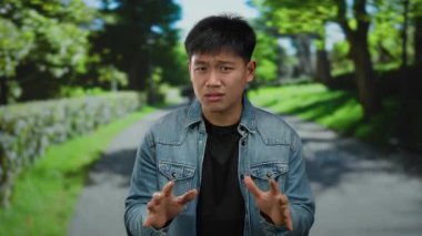 Genç adam, güneşli bir günde açık bir arka planda, yemyeşil bir parkta anlamlı jestler yapar.