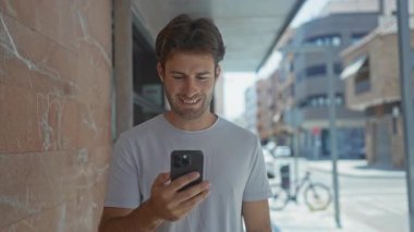 Sokakta akıllı telefonu tutan adam, ekranda gülümserken mermer duvara yaslanıyor ve mesajları izliyor; gündelik memnuniyet.