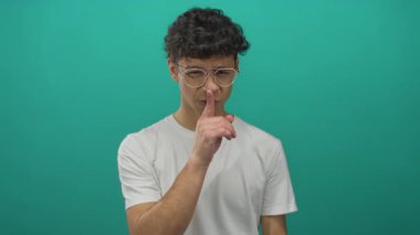 Kıvırcık saçlı, gözlüklü ve beyaz gömlekli İspanyol bir adam stüdyoda sessizlik için dudaklarını parmaklıyor..