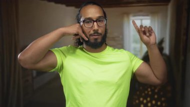 Neon yeşil tişörtlü bir adam, bir oturma odasında çerçevelenmiş bir binanın içinde el ve işaret ile bir telefon görüşmesi yapar; oyuncu bir bağlantı.