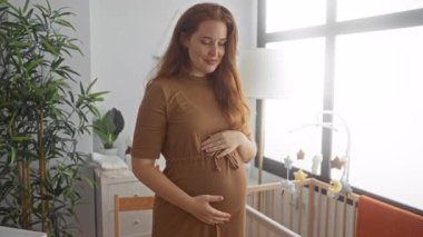 Kahverengi saçlı hamile kadın, pencerenin yanındaki ahşap karyolada karnını nazikçe tutuyor..