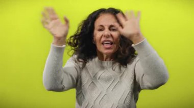 Sarı arka planda kıvırcık saçlarıyla el kol hareketi yapıp bağıran, duygularını canlı bir şekilde ifade eden, kazak giyen, hareketlerini vurgulayan parlak bir duvarın üzerine kurulmuş bir kadın..