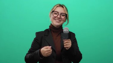 İzole edilmiş yeşil arka plan duvarına karşı mikrofonla el kol hareketi yapan bir kadın muhabir stüdyo ortamında canlı bir iletişim sahnesi sergiliyor.