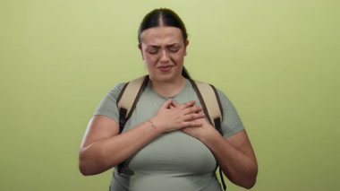 Sırt çantasıyla göğsünü tutan kıvrımlı genç bir kadın izole edilmiş sarı bir duvarın karşısında duruyor. Rahatsızlık ve endişeyi ifade ediyor. Duygularını ve gerginliğini ifade ediyor..