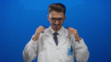 Mavi stüdyoda steteskopu ayarlarken gülümseyen gözlük takan doktor; profesyonel özgüven sağlığı.