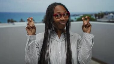 Kıvırcık saçlı bir kadın deniz kenarında gezinirken tırnak işareti yapıyor. Plaj zemininde gözlük takıyor ve bulutlu bir gökyüzünün altında rahat bir ifade takınıyor..