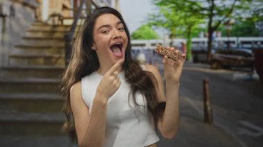 Latin kökenli genç bir kadın şehir sokaklarında ağzı açık gülümseyen granola barını işaret ediyor..