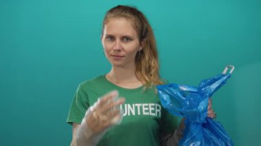 Kadın gönüllü olarak stüdyoyu işaret ederken çanta kaldırıyor ve eldiven giyen sarışın genç aktivist yerden plastik mavi çöp topluyor..