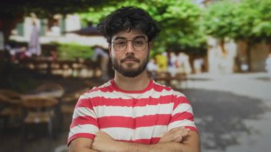 Gözlük takan sakallı genç adam güvenli bir şekilde ayakta duruyor. Arka planı bulanık, hareketli ve benzersiz bir sokak manzarası ve eşsiz bir stili olan hareketli bir sokakta kesişiyor..