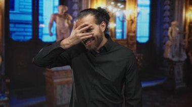 Elle yüzünü örten ve mermer heykellerin yanında süslü müze galerisinde gülümseyen adam, siyah gömlek ve pantolon askısı takıyor; eğlence özel anı.