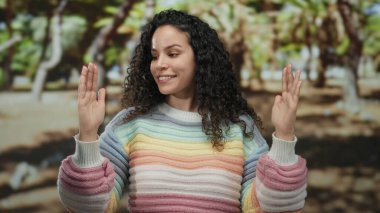 Parkta, açık havada, renkli bir süveter giymiş gözleri kapalı gülümseyen, ağaçlarla çevrili, güneşli bir ortamda el hareketi yapan bir kadın..