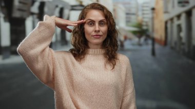 Genç bir kadın, şehir sokaklarında rahat bir süveter giyerek, modern bir yaşam tarzı için canlı ama sakin bir atmosfer izlenimi uyandıran sıradan bir selam veriyor..