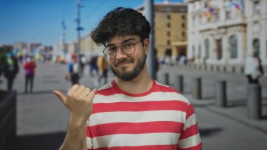 Sakallı, gözlüklü ve çizgili tişörtlü genç İspanyol adam güneşli bir günde açık havada başparmak uçlarını gösteriyor..