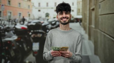 Kentsel sokak ortamında gülümseyen ve arka planda park edilmiş motosikletleri olan Norveç kronu banknotları tutan adam..