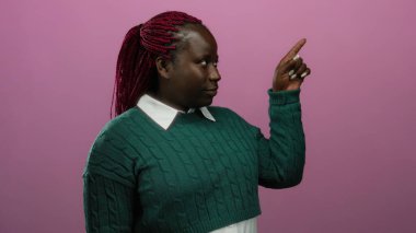Pembe arka planda örülü saçları olan, yeşil kazaklı bir kadın stüdyo ortamında yön ya da ilgi gösteren bir şeyi işaret ediyor..