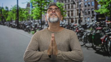 Elleri dua jestinde birbirine bastırılmış bir adam, gözlük takan, çıplak elleri görünen, park halindeki bisikletlerin yanındaki bir Amsterdam caddesine bakan; umut ışığı..