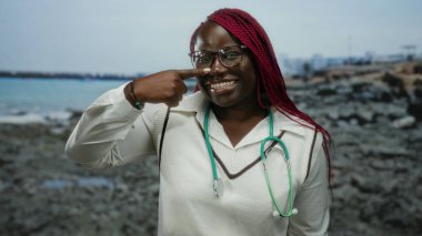 Saçları örgülü ve steteskoplu gülümseyen kadın kayalık bir sahile burnunu işaret ederek arka planda sakin mavi bir deniz olan neşeli bir atmosfer yaratıyor..