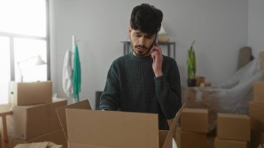 Genç adam yeni evindeki kutuları açarken telefonla konuşuyor. Etrafı taşınan kutular ve doğal ışıklarla çevrili. Geçiş ve modern yaşam hissini aktarıyor..