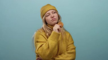 Sarı kazaklı ve şapkalı kadın mavi arka planda kurabiye tutuyor. Düşünceli ve meraklı görünüyor. Stüdyo ortamında çekilmiş genç ve rahat bir ifade..