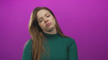 Yeşil kazaklı genç kadın başını eğiyor ve elleri mor stüdyo ortamında ağzını kapatıyor..