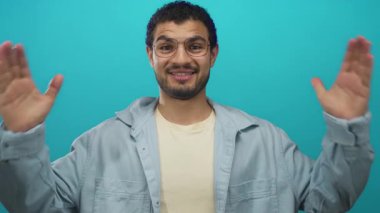 Gözlüklü sakallı adam stüdyoda turkuaz duvara karşı hafif gömlek giyerek el ele tutuşuyor; iyimserlik..