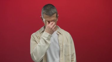 İzole edilmiş kırmızı arka planda yüzü olan bir adam stresli ya da endişeli, gözleri kapalı sıradan bir gömlek giyiyor..