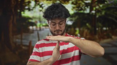 Sakallı, gözlüklü, çizgili tişörtlü, dışarıda yeşil bir parkta mola veren İspanyol bir genç..