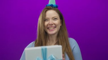Stüdyoda elinde mavi kurdeleyle bağlı büyük beyaz bir hediye kutusu tutan kadın canlı mor arka planlı parti şapkası takıyor..