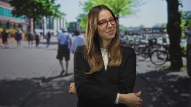 Kırmızı gözlüklü kadın park halindeki bisiklet ve yayaların yanında kollarını kavuşturuyor; kendine güveni olan profesyonel bir kadın..