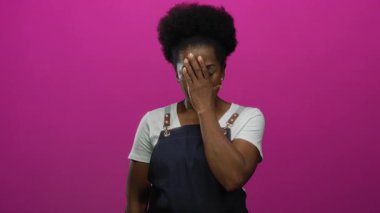 Önlüklü kadının elleri kapalı pembe arka planı duygusal ifade ya da strese işaret ediyor.