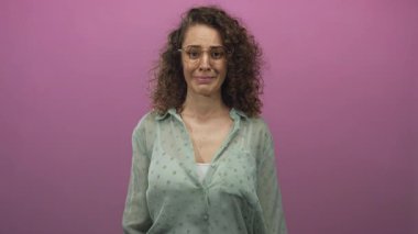 Stüdyoda pembe duvara karşı parmak ve buruşukluklar gösteren bir kadın, gözlüklü ve kıvırcık saçlı yeşil benekli bluz giyiyor; hayal kırıklığı.