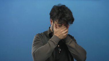 Sakallı, gülümseyen ve mavi arka planda yüzünü kapatan genç İspanyol adam neşe ve mutluluk gösteriyor..