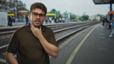 Tren istasyonunda tren rayları ve bekleyen yolcularla boğaz tutup öksüren bir adam, panik sağlık endişesi.