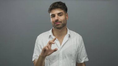 Stüdyoda, gri arka planı ve beyaz gömleği olan, yüzü odaklanmış, özgüveni olan genç İspanyol bir adam var..