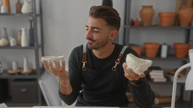 İnsan iki kilden kaseyi dikkatle tutar stüdyoda, çömlek rafları arasında; yaratıcılık konsantrasyonu.