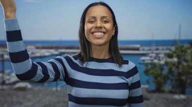 Güneşli bir günde deniz kenarında gülümseyen bir kadın çizgili bir gömlek giyiyor, etrafı tekneler ve canlı mavi suyla çevrili, neşeli, rahat bir yaz havası sergiliyor..