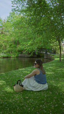 Sıcak bir yaz gününde yapraklı ağaçların altında saman torbası taşıyan, yapraklı bahçedeki sakin bir havuzun kenarında yemyeşil çimlerde oturan bir kadın.