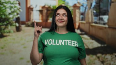 Yeşil gönüllü tişörtü giymiş iri yarı bir kadın güneşli park ortamında yukarıya doğru bakıyor. Açık havada gönüllülük ve toplum ruhunu sergiliyor..