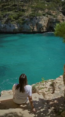 Santanyi, Mallorca 'daki kayalık bir sahilde turkuaz sularda dinlenen bir kadın Akdeniz güneşinin ve manzaranın tadını çıkarıyor..