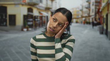 Şehirde hareketli bir sokakta düşünceli görünen genç bir kadın ifadeleriyle duygularını sergiliyor, açık havada çizgili bir gömlek giyiyor..