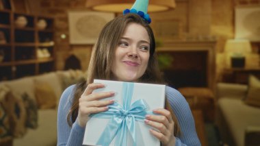 Mavi kurdeleli bir hediye kutusu tutan ve rahat oturma odasında gülümserken parti şapkası takan bir kadın..