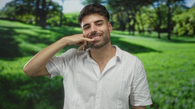 Gülümseyen adam, yeşil güneşli parkta elini yüzüne yaklaştırarak çağrı hareketi yapar. İletişim mutluluğu..