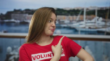 Kırmızı gömlekli genç kadın, liman korkuluklarının yanındaki yelkenlileri işaret ediyor; kamu hizmeti şüphesi.