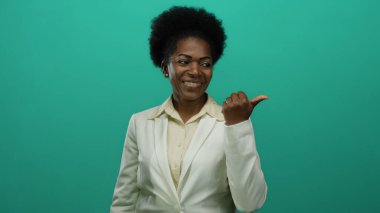 Beyaz takım elbiseli, afro saçlı bir kadın yeşil bir arkaplanı işaret ederek, dostça bir gülümsemeyle onaylama ve pozitifliği dile getiriyor..