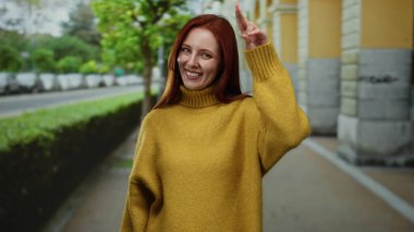 Kırmızı saçlı bir kadın şehir caddesinde sıcacık gülümsüyor, sarı bir süveter giyiyor, şehir yeşilliği ve mimarisi ile çevrili, neşeli bir atmosferi olan canlı bir açık hava manzarası yaratıyor..